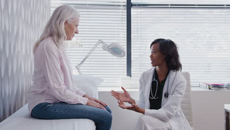 Mature-Woman-In-Consultation-With-Female-Doctor-Sitting-On-Examination-Couch-In-Office