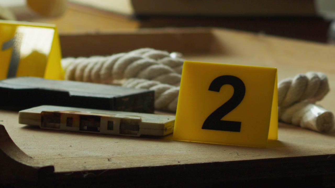 Slow-motion close-up of a marker placed near rope and a cassette tape, suggesting layered clues in a forensic investigation scene.
