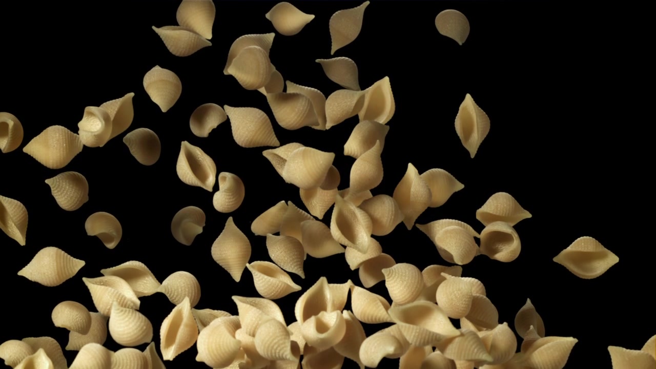Trockene Conchiglie-Nudeln fallen in Zeitlupe vor schwarzem Hintergrund.
