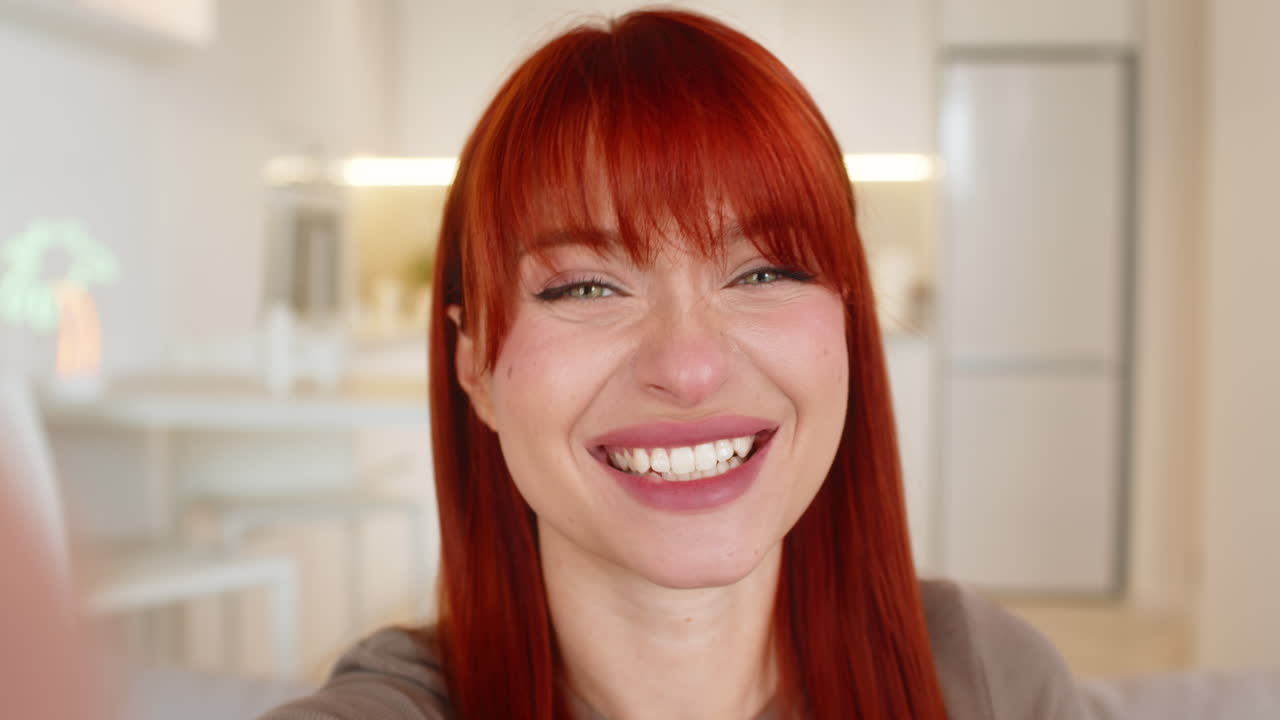 Giovane donna che mostra uno smartphone alla fotocamera, saluta con un cenno caloroso e un sorriso gentile durante una videochiamata POV