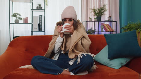 Sick-young-woman-wear-hat-wrapped-in-plaid-sit-alone-shivering-from-cold-on-sofa-drinking-hot-tea