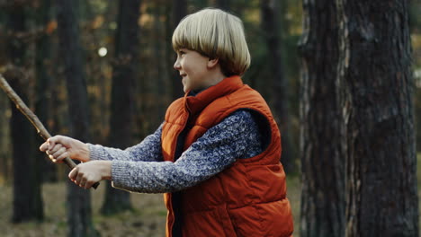 lindo niño rubio caucásico jugando con un palo en el bosque alegremente