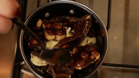 Eine-Zerbrochene-Tafel-Milchschokolade-Mit-Haselnüssen-Und-Butter-Wird-Auf-Dem-Gasherd-Geschmolzen,-Während-Eine-Frau-Mit-Ehering-In-Einem-Kleinen-Topf-Zum-Schmelzen-Rührt