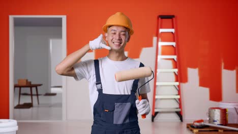 pintor hombre asiático con casco de seguridad sonriendo y haciendo llamarme gesto a la cámara mientras está de pie con la pared de pintura en la habitación