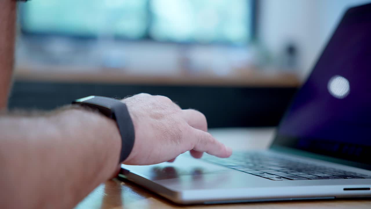 Vista di profilo di braccia e mani che digitano su un computer portatile. Il soggetto indossa un fitness tracker al polso mentre lavora. Lo sfondo sfocato suggerisce un ambiente interno luminoso
