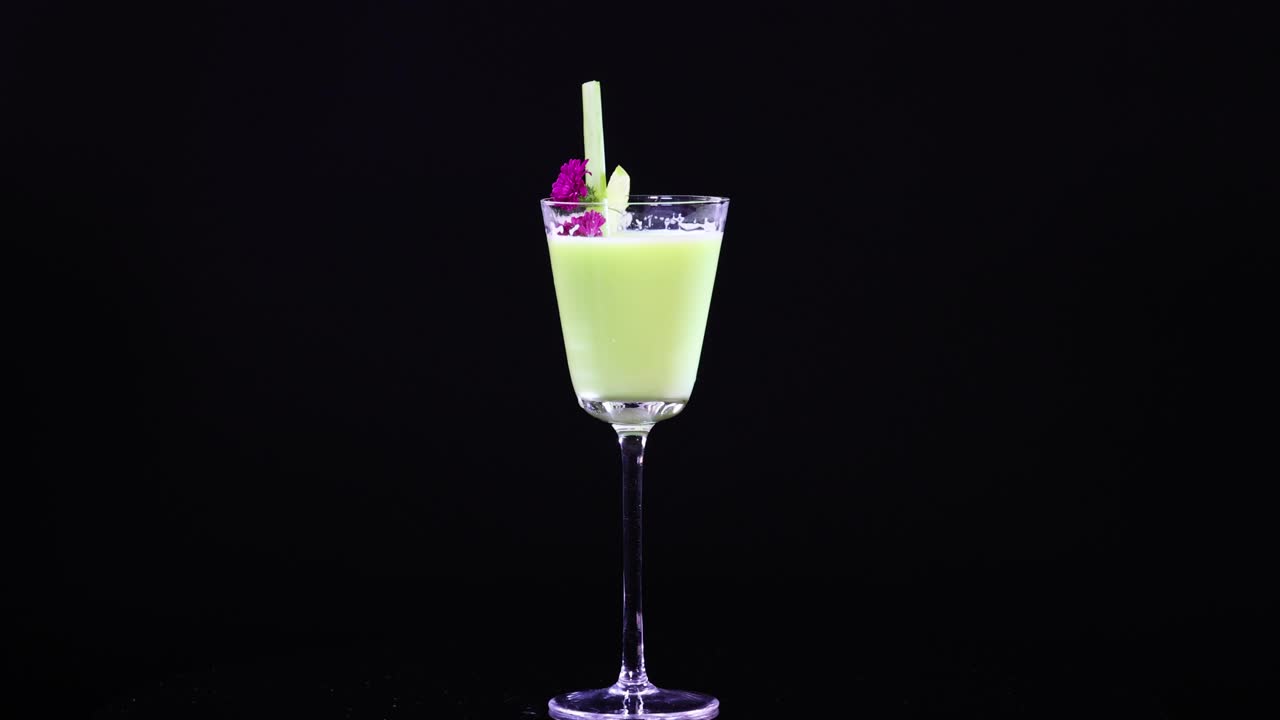 A vibrant green cocktail in a glass with garnish, set against a black background. Bright lighting highlights the drink's vivid color