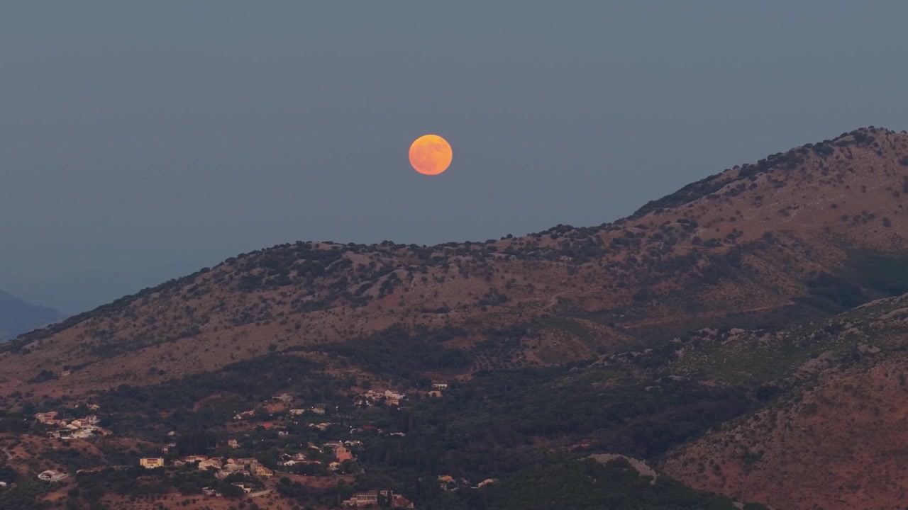 A breathtaking time-lapse video of a vibrant, orange full moon rising high over the mountain ridges of Corfu Island, Greece. The fading twilight sky perfectly contrasts with the bright lunar surface,