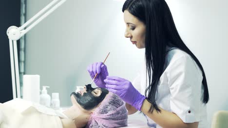 attractive beautician in medical coat and blue gloves applying black cosmetic mask on face to a woman in beauty salon. health and skin care.