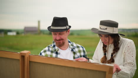 an illustrator in a checkered shirt smiles as he works on his painting, set against a lush green landscape. beside him, a woman in a white dress and hat stands, observing his work with interest