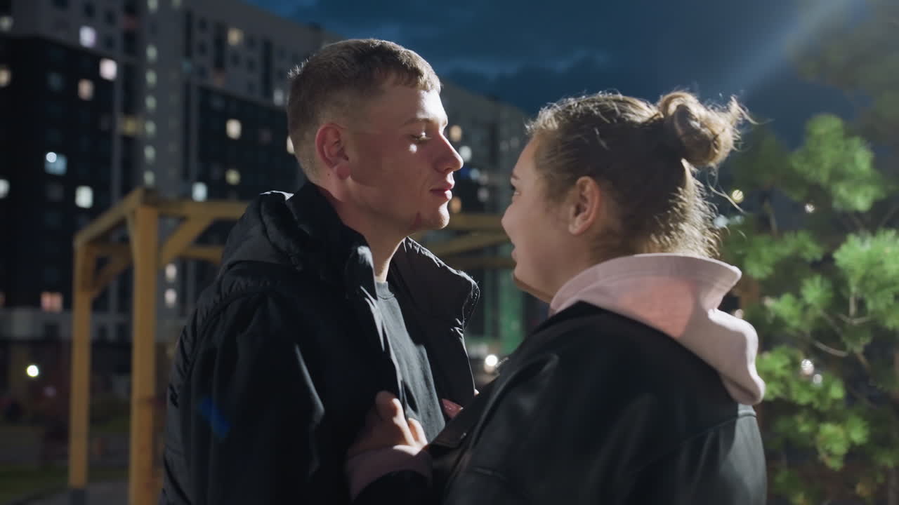 White man gives his lady gentle peck on forehead while she holds onto his jacket affectionately under evening sky with soft city lights and tall buildings