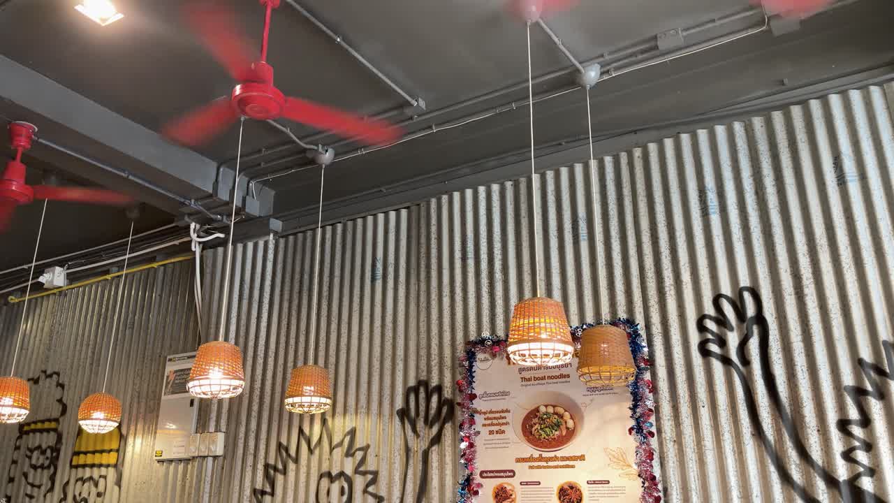 Static upward view of spinning red ceiling fans and warm hanging lights in corrugated noodle shop