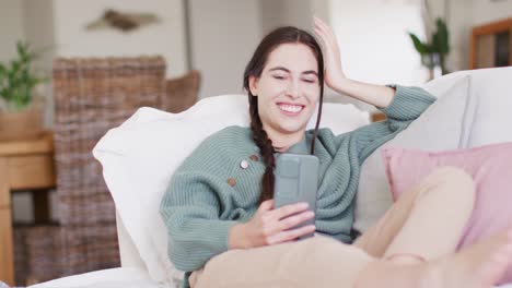 mujer caucásica feliz sentada en el sofá y usando un teléfono inteligente