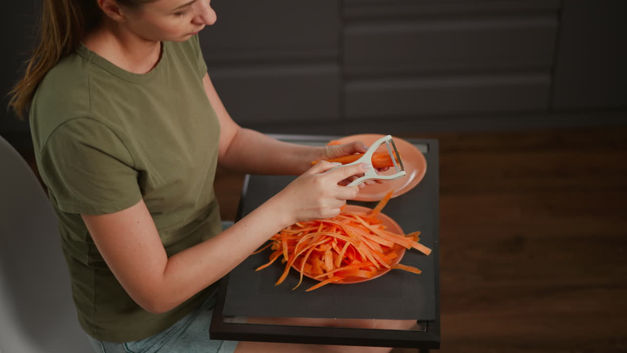 Top down view of woman in green shirt preparing carrot with white peeler, long orange strips filling plate on black table, dark kitchen cabinet and wooden floor in background of cooking setup