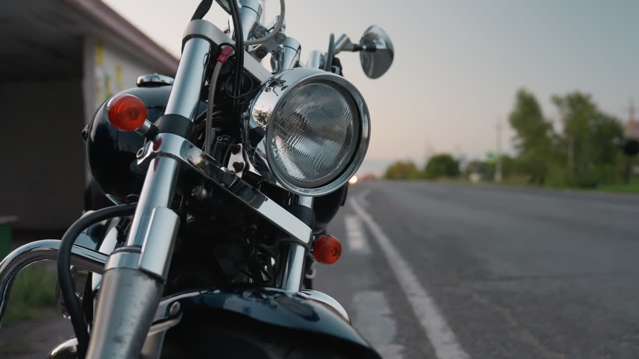 Close up parked motorcycle with chrome details shining under evening light while car passes on nearby lane and blurred headlamp glows in distance, creating atmosphere of travel