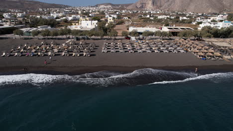 santorini island, greece, aerial view of black sand beach, parasols, sunbeds and aegean sea waves, revealing drone shot