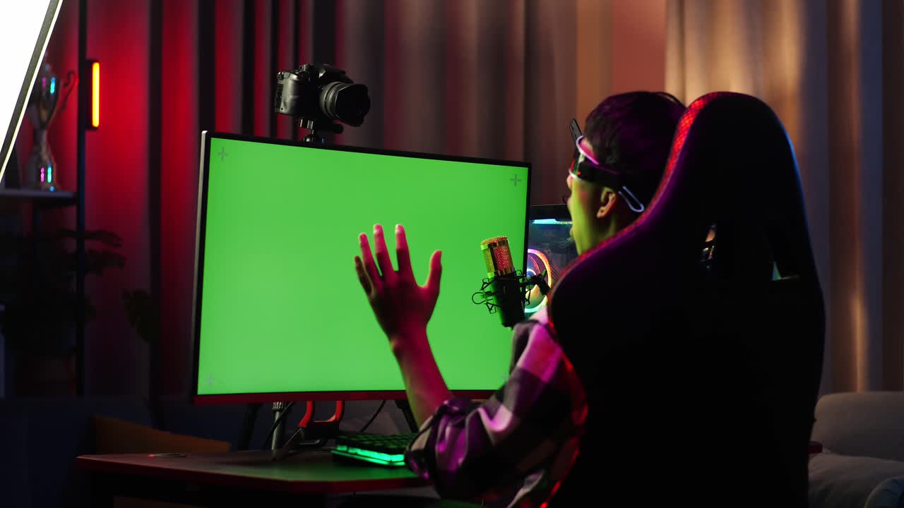 Back View Of Asian Boy Streamer Playing Game, Touching, And Swiping Experience Virtual Reality In Futuristic Goggles. Green Screen Personal Computer On Desk Illuminated By Rgb Led Strip Light
