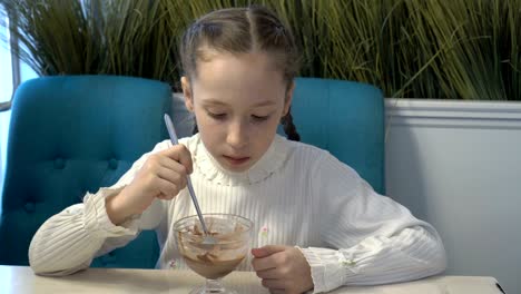 una niña con un vestido blanco, con colas, se sienta y come un dulce postre-helado en un café. está comiendo con una cuchara larga de una taza de vidrio. retrato.