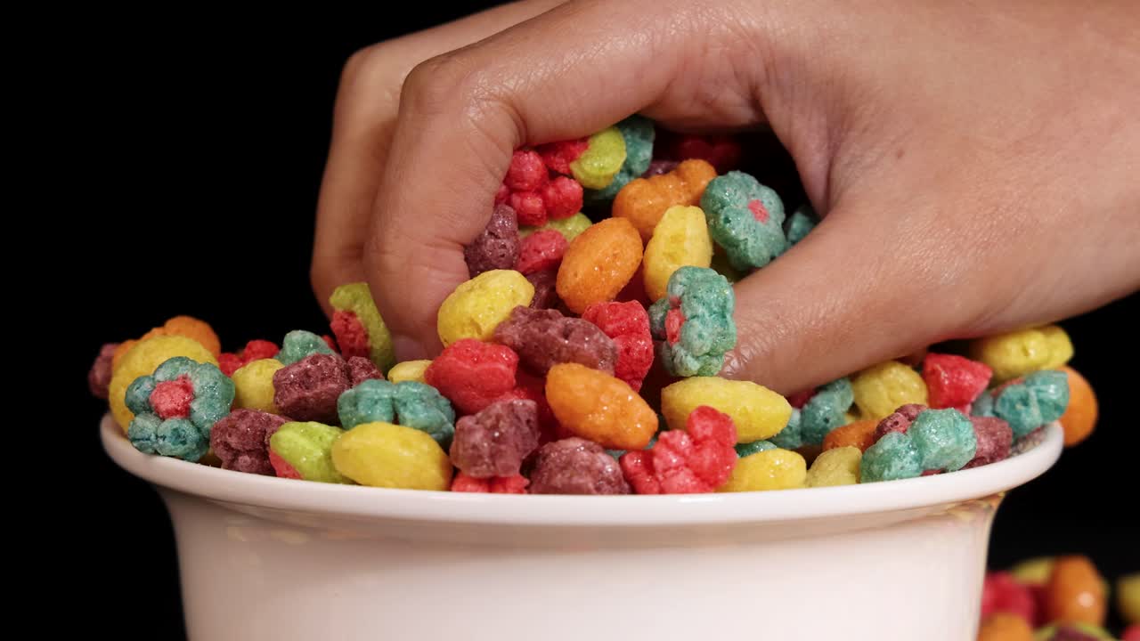 A hand interacts with vibrant cereal in a bowl against a black background, showcasing dynamic movement and colorful visuals