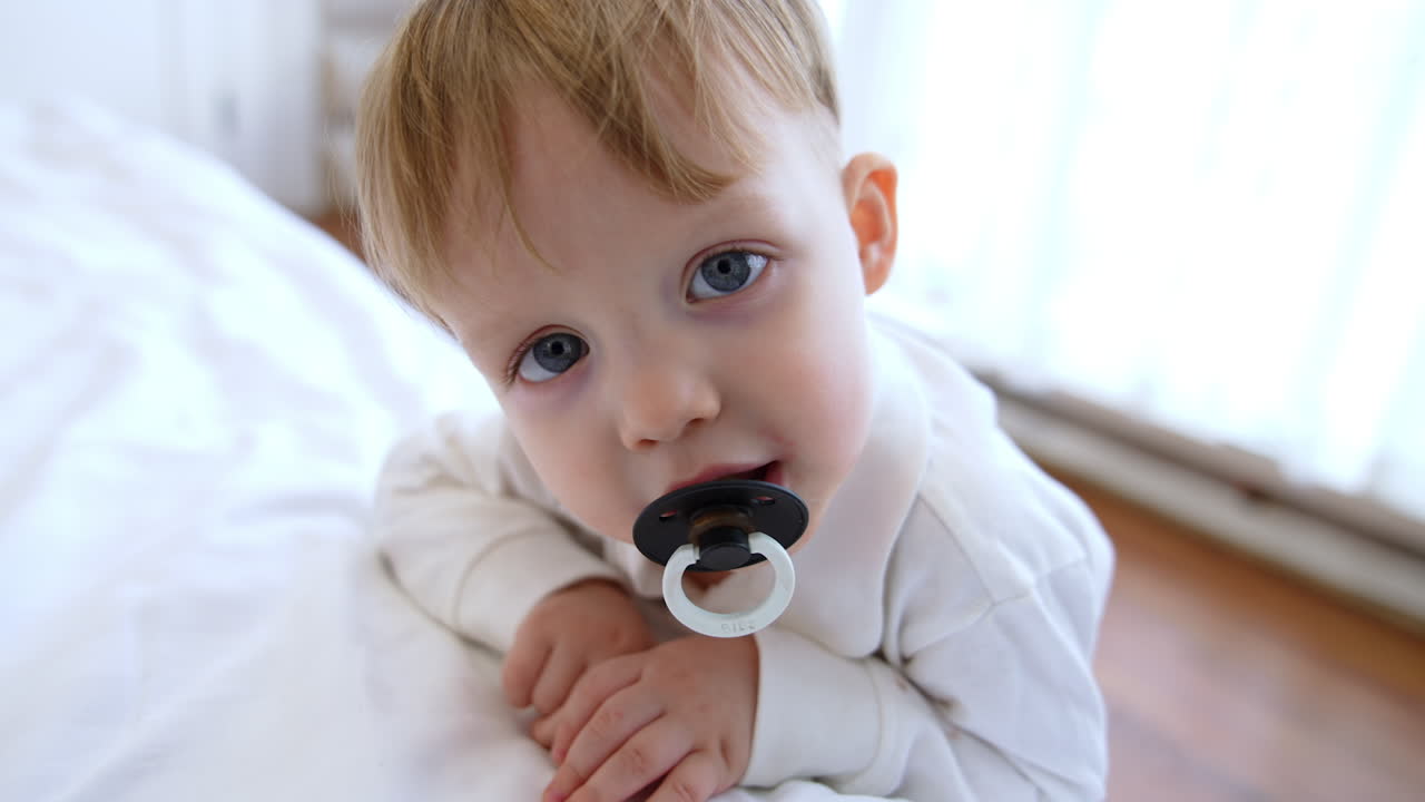 Sweet Caucasian toddler with pacifier stands smiling at the bed. Close up. Woman sits on the floor at backdrop in blur.