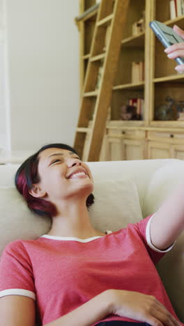 vertical video of biracial teenager girl sitting on sofa and using smartphone, in slow motion