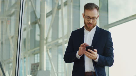 Portrait-Shot-Of-The-Young-Handsome-Businessman-Taping-And-Texting-On-The-Smartphone-In-His-Hands,-Then-Rising-Head-And-Smiling-To-The-Camera