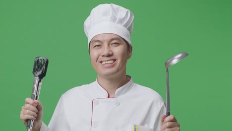 Close Up Of Asian Man Chef Smiling And Showing Tongs And Ladle To Camera While Standing In The Green Screen Background Studio
