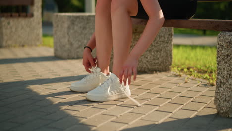 partial view of lady sitting on bench loosening shoelace of white sneakers on right leg while holding shoelace with right hand, with greenery in background and stones