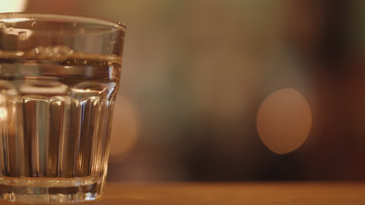 A clear glass of water is lifted from and returned to a wooden table in a dimly lit restaurant, with warm bokeh lights in the background