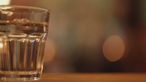 a clear glass of water is lifted from and returned to a wooden table in a dimly lit restaurant, with warm bokeh lights in the background