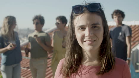 retrato de una mujer caucásica con gafas de sol en la cabeza mirando a la cámara y sonriendo mientras tiene una fiesta en la azotea con amigos