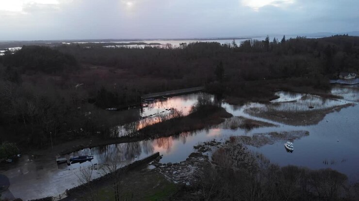 Una suggestiva veduta aerea cinematografica della costa di Lough Corrib a Cong, Contea di Mayo, con acque calme, piccole imbarcazioni e un drammatico riflesso del tramonto. Perfetto per documentari di viaggio e natura