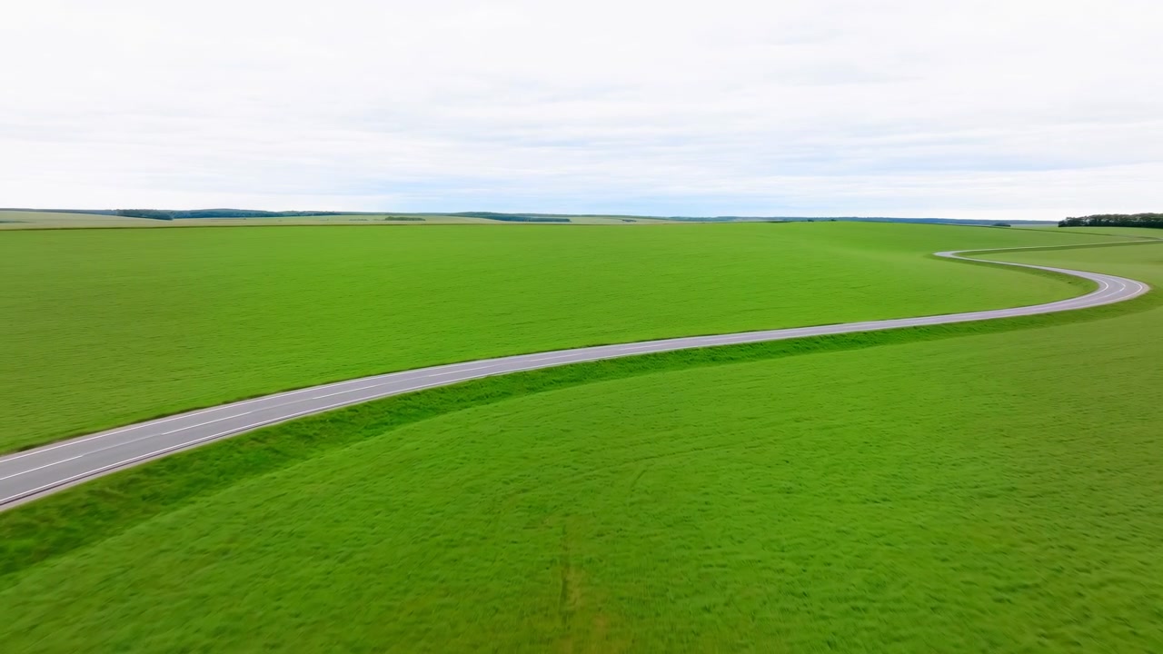 Camera moves smoothly above an expansive green field while a winding gray road shifts across the frame, with distant farmland and a bright clouded horizon completing the open rural view.