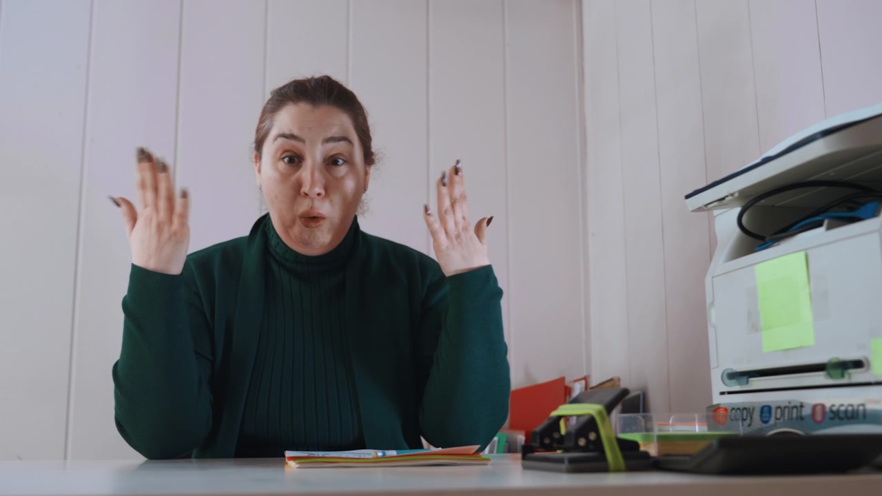 Caucasian woman at desk in home office, studying paperwork and gesturing to throat, consulting telehealth, printer and computer visible, tense expression, attempting to describe symptoms and prepare