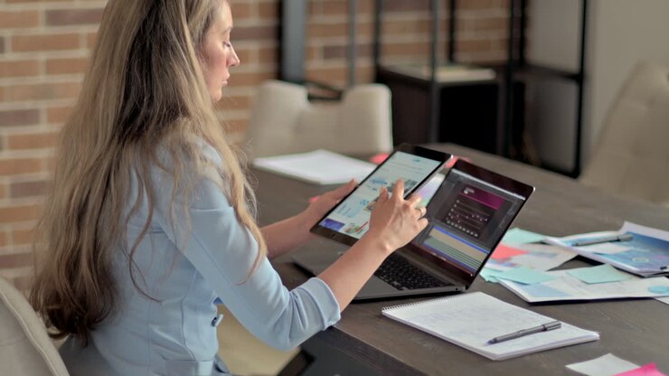 Woman in blue blazer working on a tablet in front of a laptop, at a desk, in an office