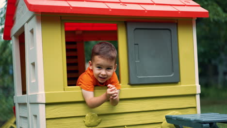 lovely smiling toddler boy peeps out of the window in the playhouse. happy child playing outdoors in summer at the playground.