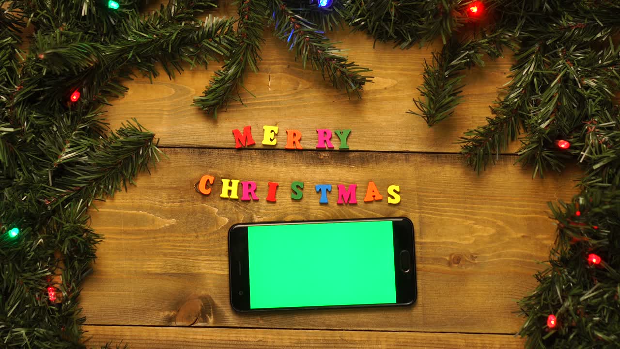 teléfono con pantalla verde y la inscripción feliz navidad en la mesa de madera con guirnaldas parpadeantes.