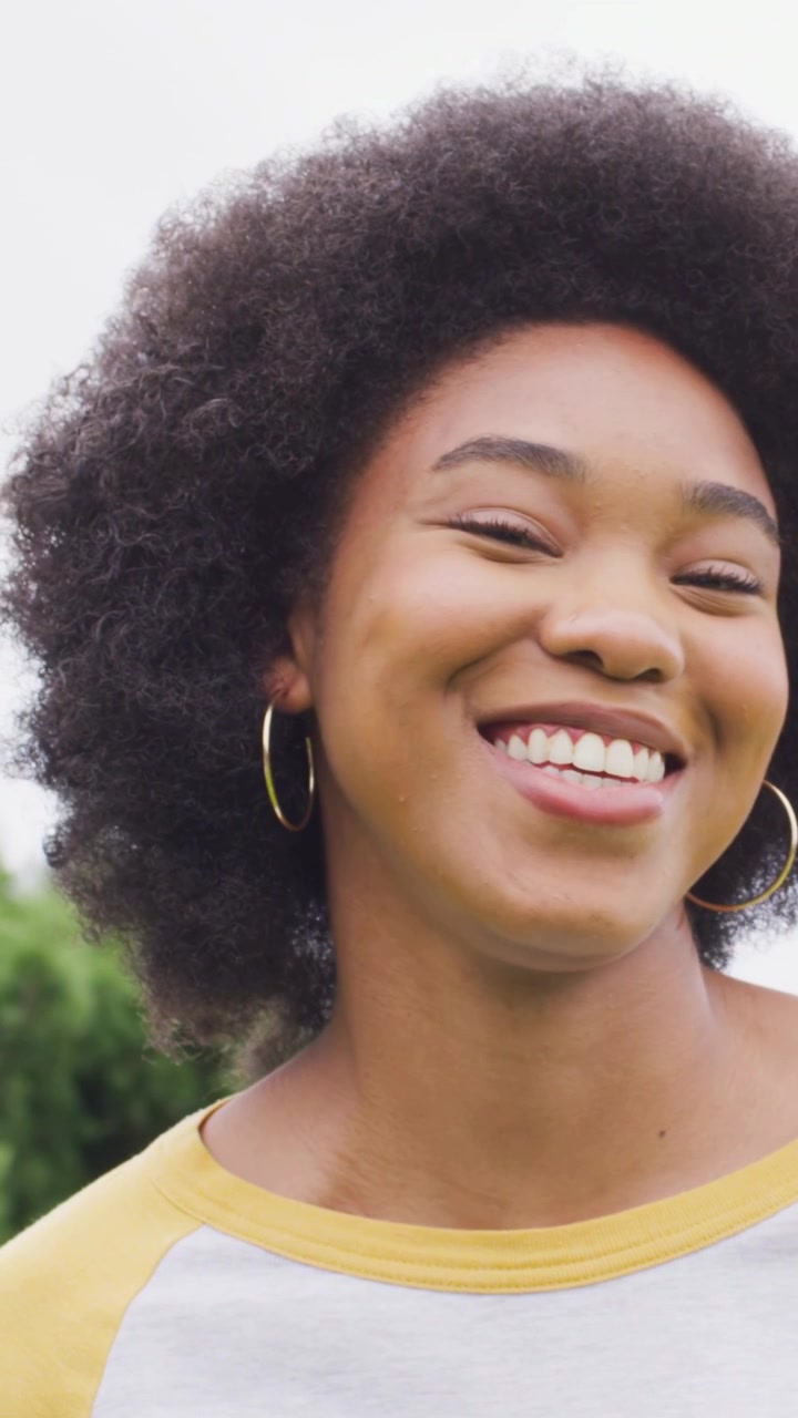 Vídeo vertical: Mulher sorrindo com blusa branca e argola dourada, mudando o olhar e respondendo à câmera no jardim.