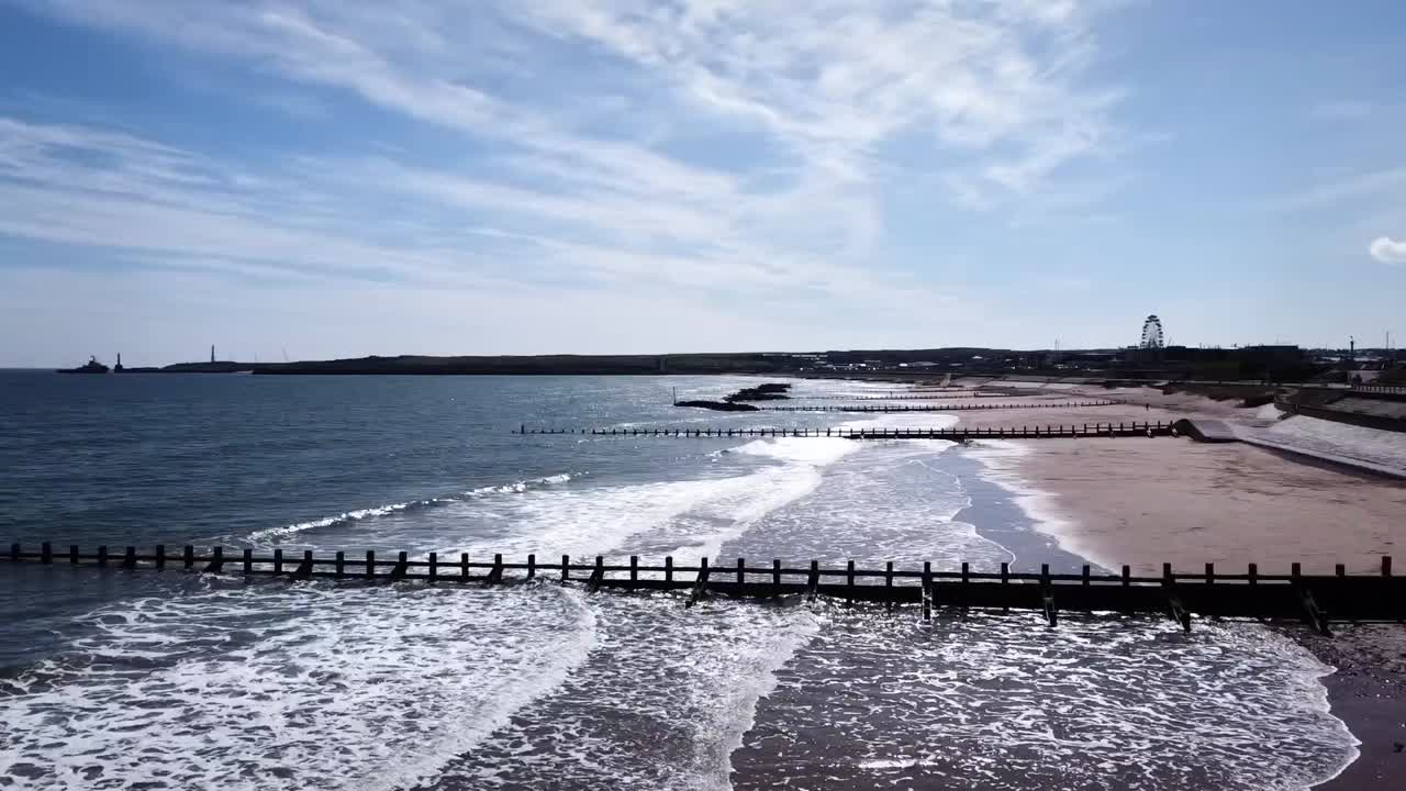 Waves gently lap against a sandy Scottish shore as a wooden groyne stretches into the North Sea. Under a bright blue sky, distant lighthouses stand along the calm, picturesque coastline.