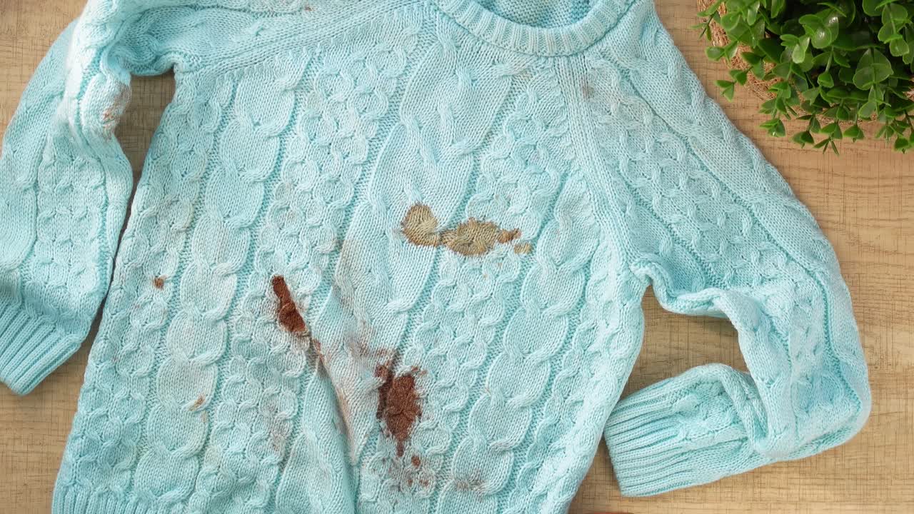 Hands holding a dirty light blue cable-knit sweater with brown stains