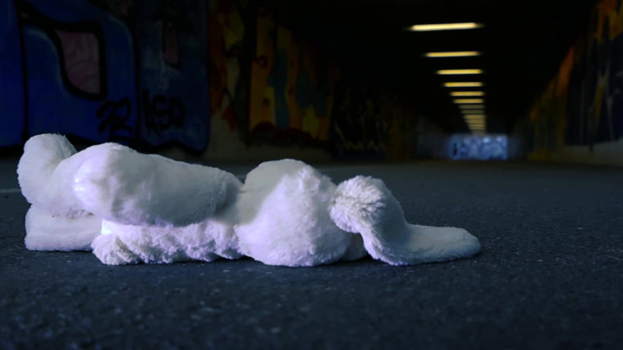 Coniglio di peluche bianco, perso e dimenticato, giace sull'asfalto di un sottopassaggio pedonale buio e coperto di graffiti, simboleggiando solitudine, abbandono e l'innocenza perduta dell'infanzia.
