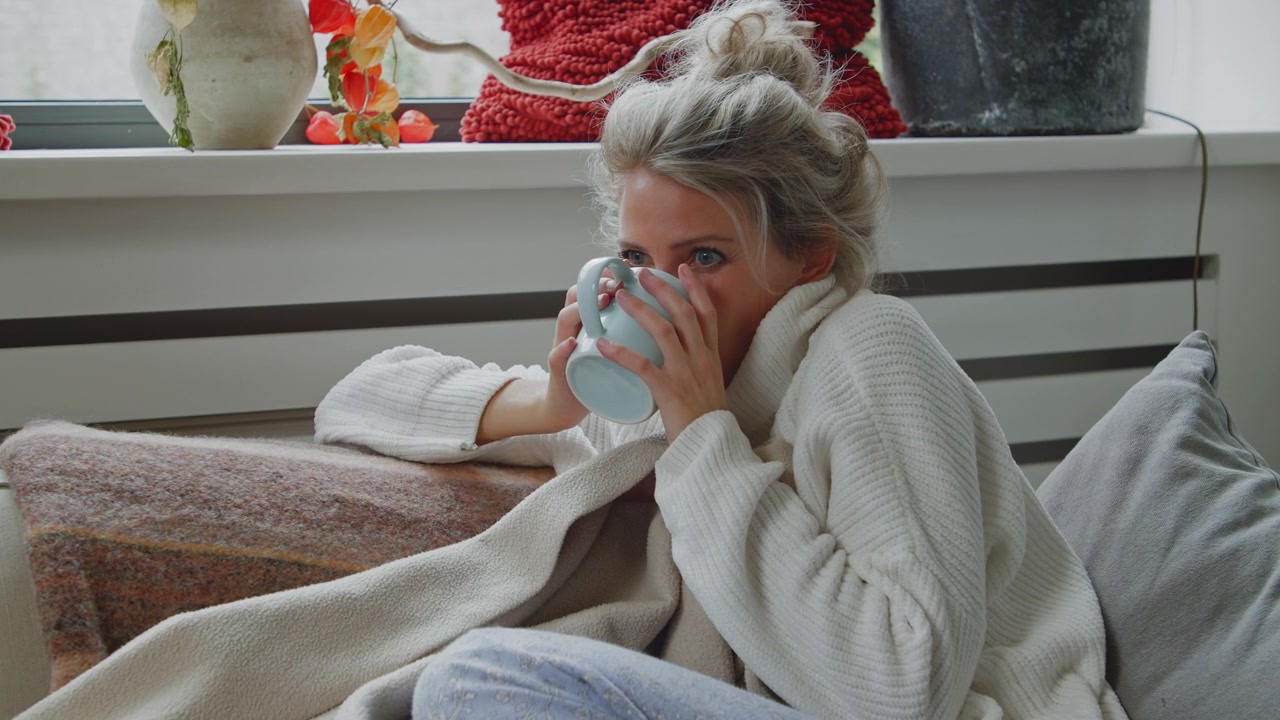 Donna infreddolita, quasi malata, che si mette comoda sotto una coperta accogliente con una tazza di tè caldo o caffè mentre cerca di sentirsi meglio.