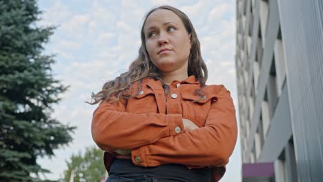 mujer en chaqueta naranja de una escena al aire libre con los brazos cruzados y una mirada pensativa inspeccionando sus alrededores
