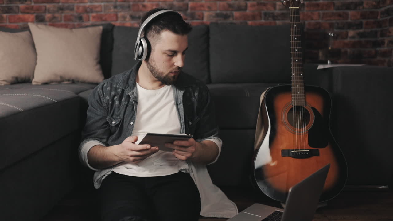 joven músico escuchando música con laptop y auriculares inalámbricos tomando notas en casa