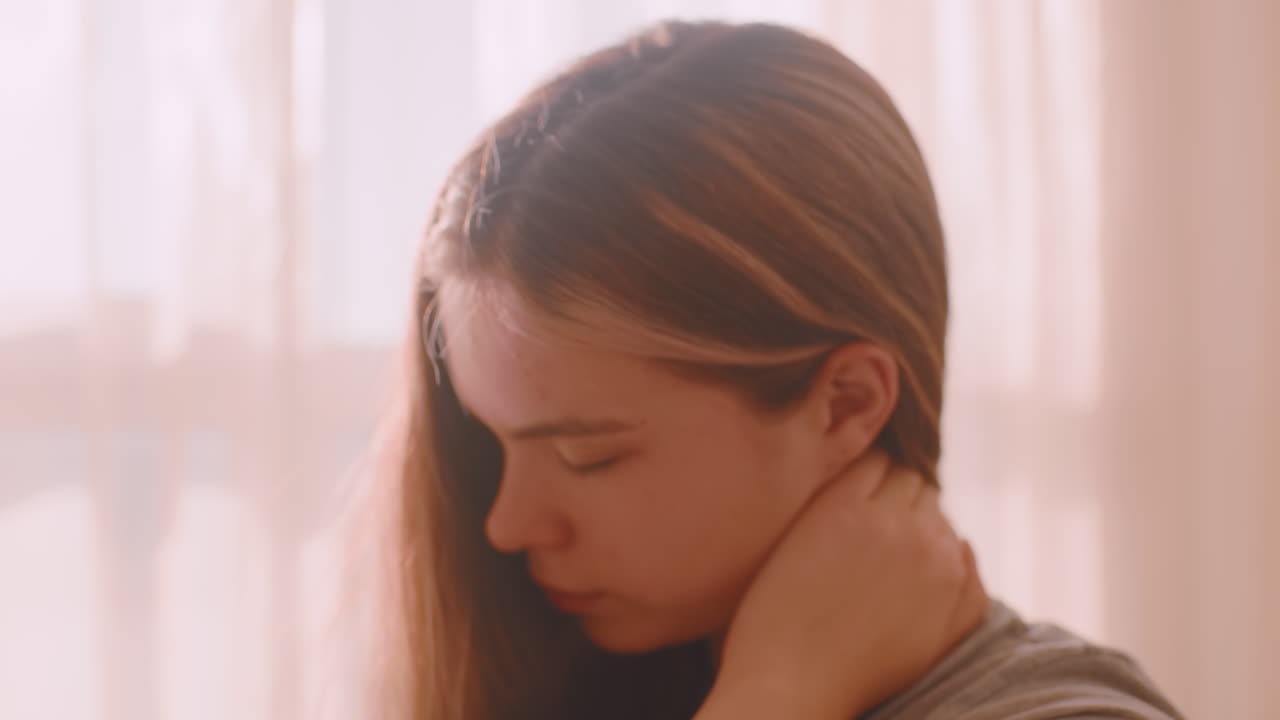 Young woman gently turns neck while seated in soft natural light near window with translucent curtain, expressing muscle tension or calm stretch
