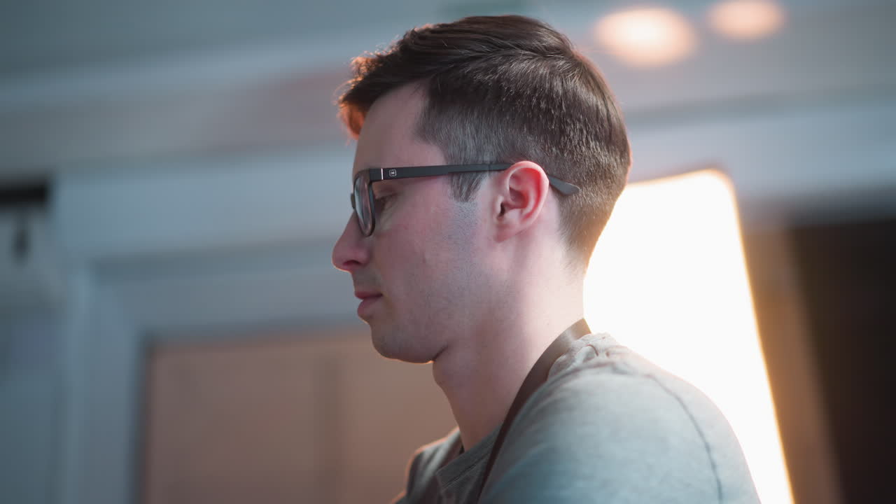Person in glasses standing in TV studio, looking at equipment, reflecting broadcast lights and graphics on glasses, with background featuring lighting setup and studio environment