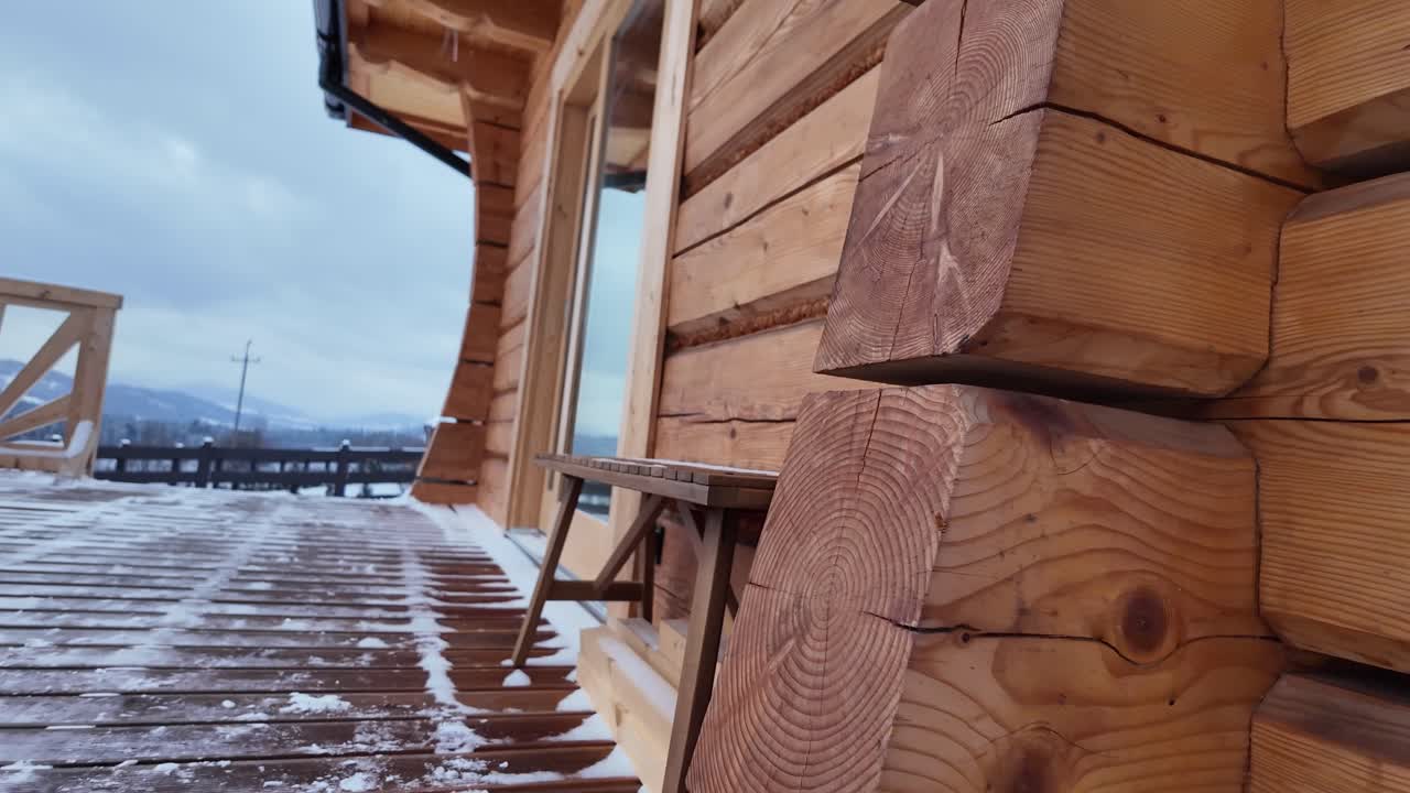 Camera glides along a wooden terrace of a log cabin during wintertime, revealing a rustic bench and serene winter mountain scenery
