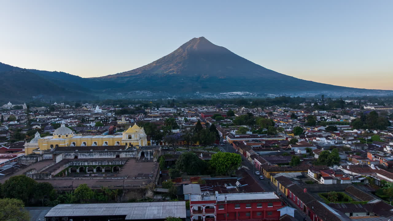 Scenic aerial view of Antigua Guatemala at sunrise, featuring colonial architecture and Volcán de Agua dominating the skyline.