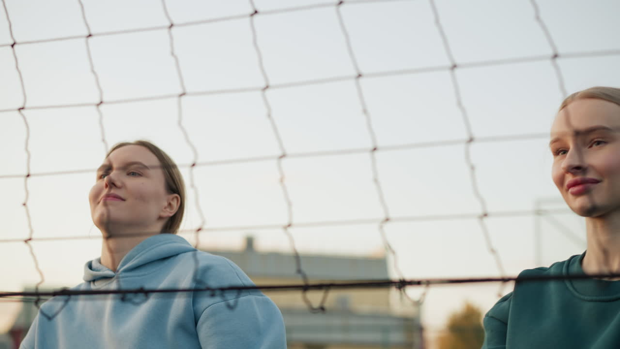 초록색과 파란색 후디를 입은 팀 동료들이 함께 점프하고, 손을 잡고 기으로 볼리볼을 망으로 돌려보내며, 야외 경기에서 배경으로 보이는 건물