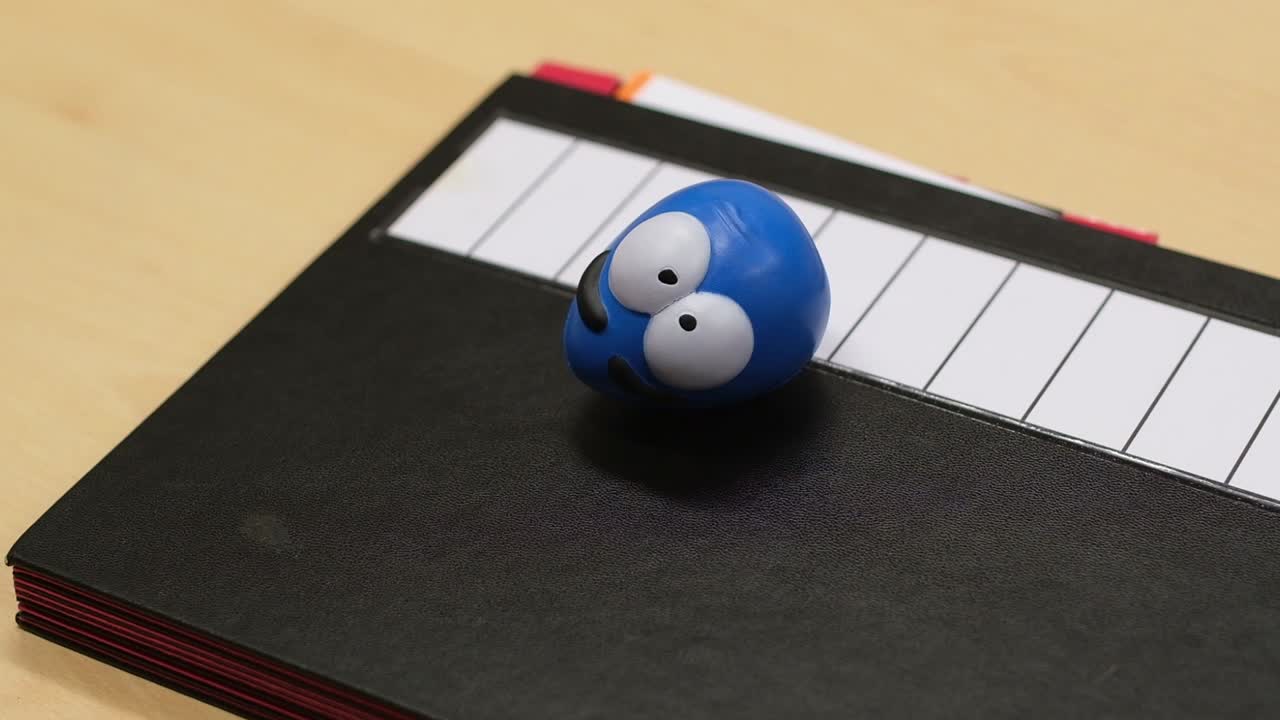 Defocus of a small toy egg with large white eyes sitting on a black paper folder in office environment. Steady camera angle