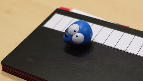 defocus of a small toy egg with large white eyes sitting on a black paper folder in office environment. steady camera angle